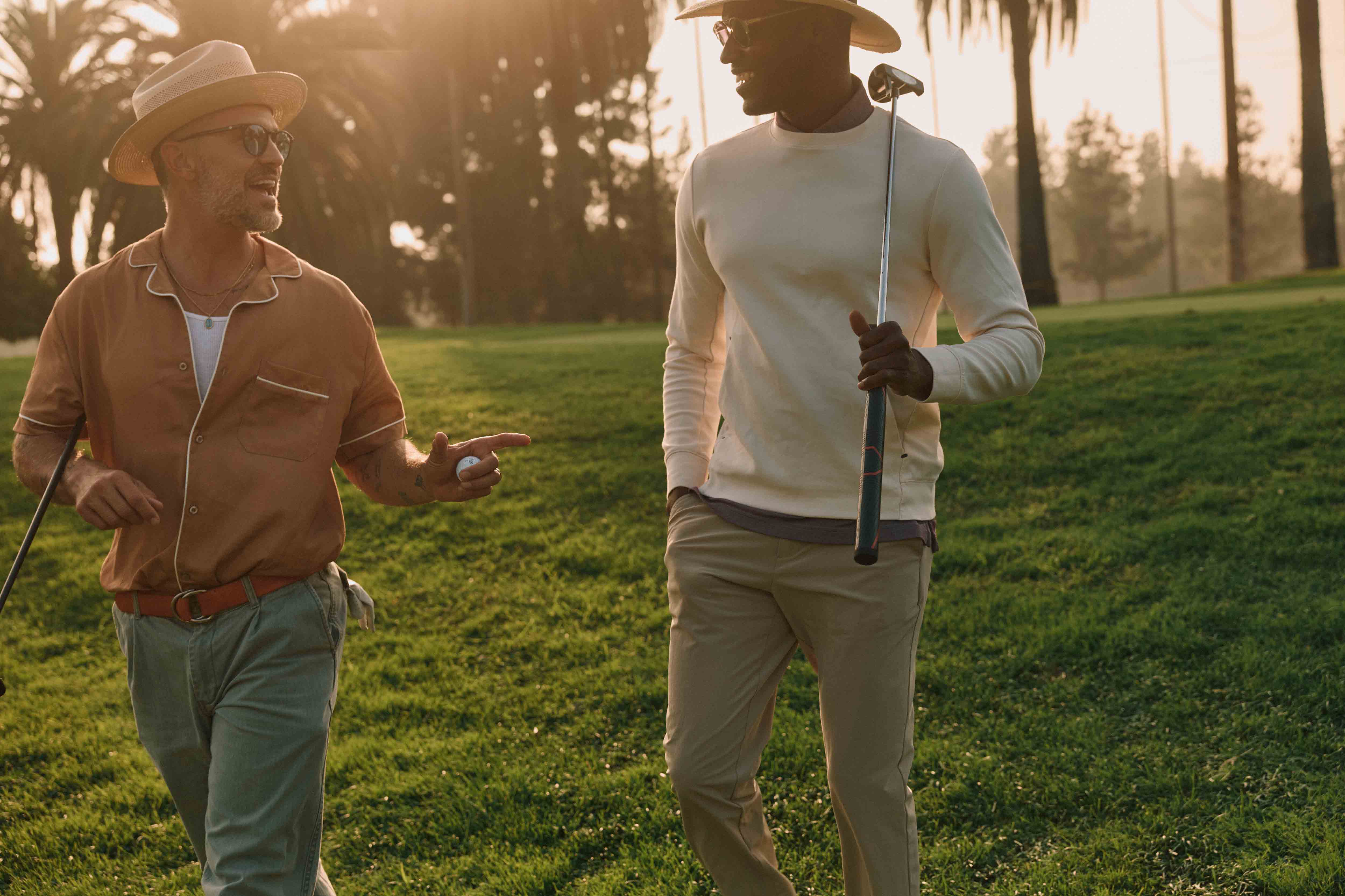 Two stylish men walk on a sunny golf course, one holding a club, the other a ball, with palm trees and green grass in the background.