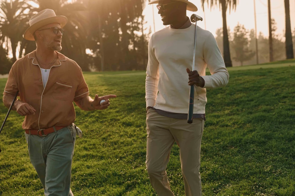 Two stylish men walk on a sunny golf course, one holding a club, the other a ball, with palm trees and green grass in the background.