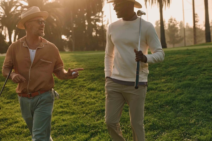 Two stylish men walk on a sunny golf course, one holding a club, the other a ball, with palm trees and green grass in the background.