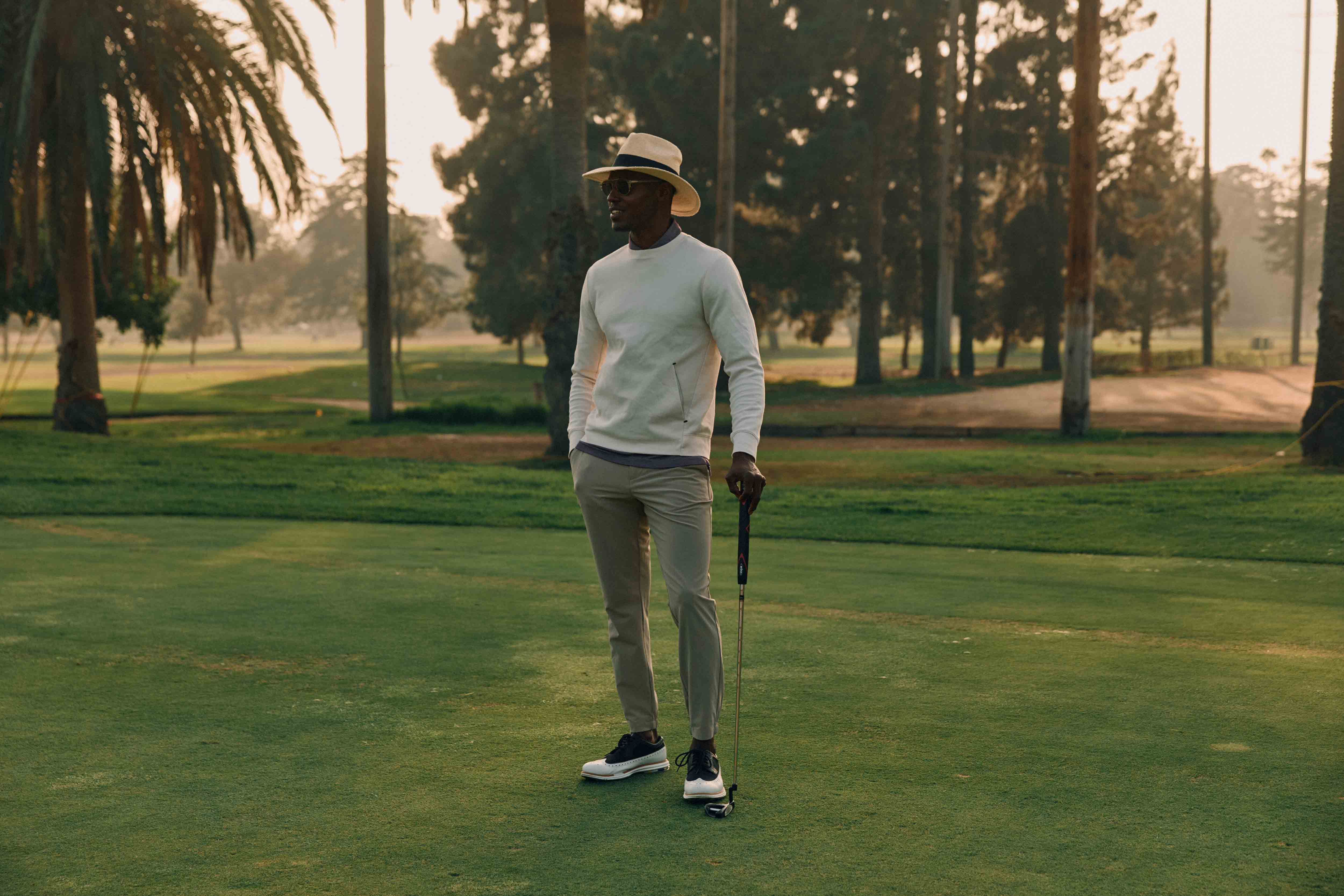An African-American man wearing a hat and holding a golf club stands on a golf course with tall trees and a green lawn.
