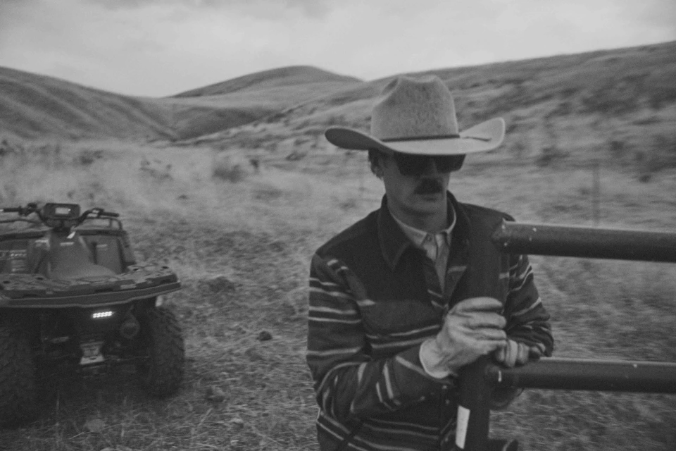 Black and white portrait of a cowboy in hat and sunglasses, resting his arms on a metal fence, with an ATV in the background.