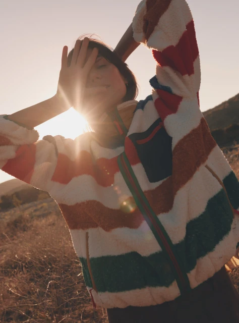 Woman in striped jacket shielding eyes from sun in rural landscape with warm light and grassy field, creating a vibrant, natural portrait.