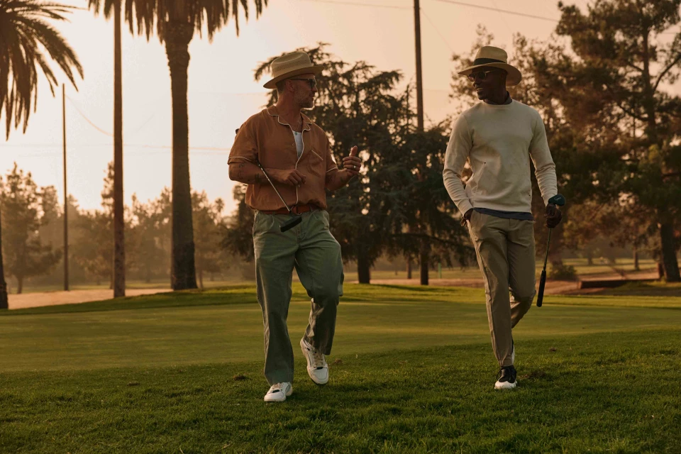 Two men walk and talk on a golf course on a sunny day, wearing stylish hats and holding golf clubs, with palm trees in background.