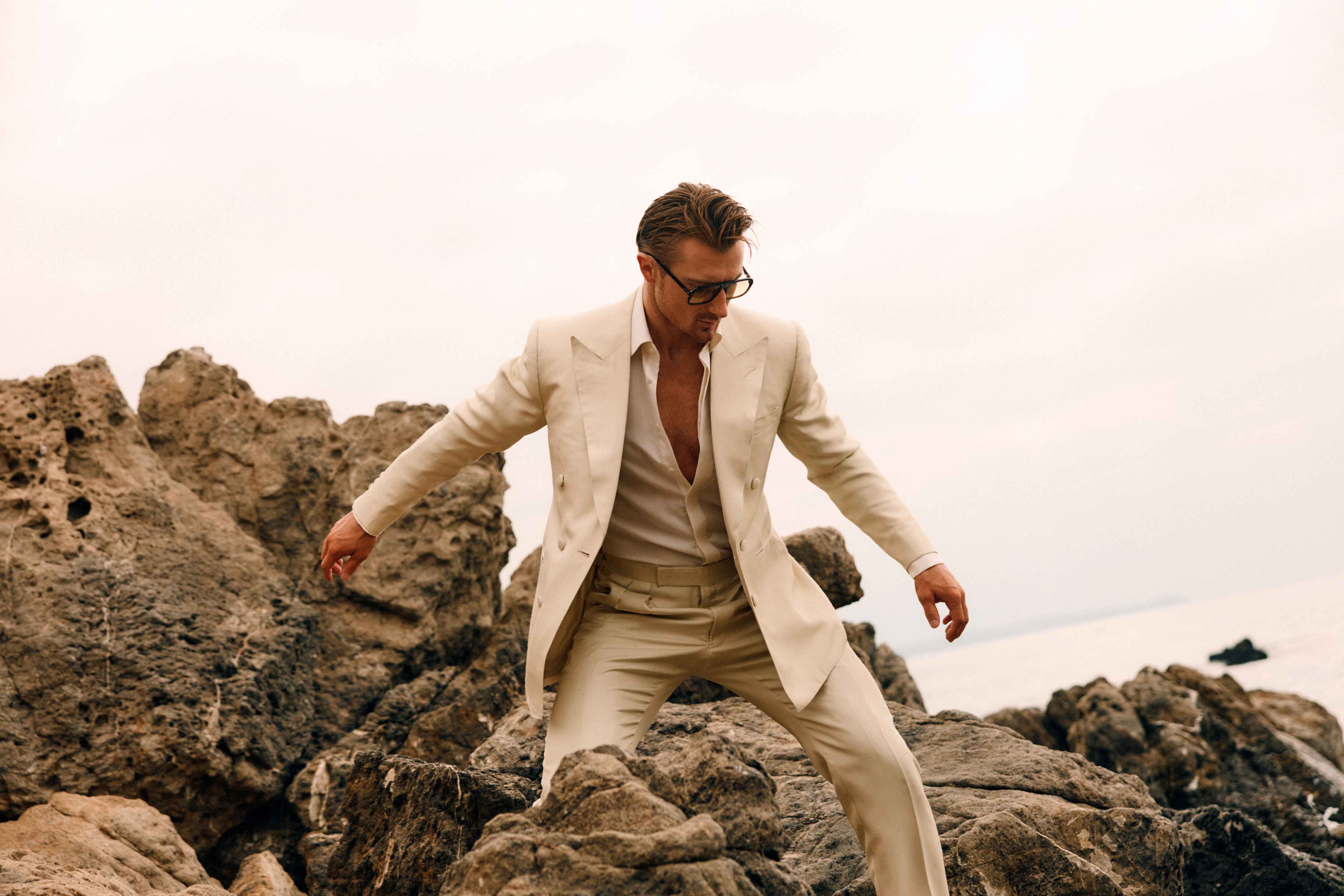 Man in a tan suit stands on rocks near the ocean. He has blond hair, wears sunglasses and an unbuttoned shirt, posing outdoors.