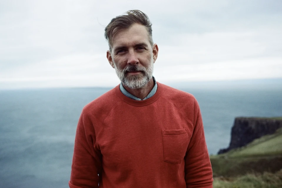 Portrait of a man with a beard wearing a red sweatshirt, standing on a grassy cliff overlooking the ocean on an overcast day.