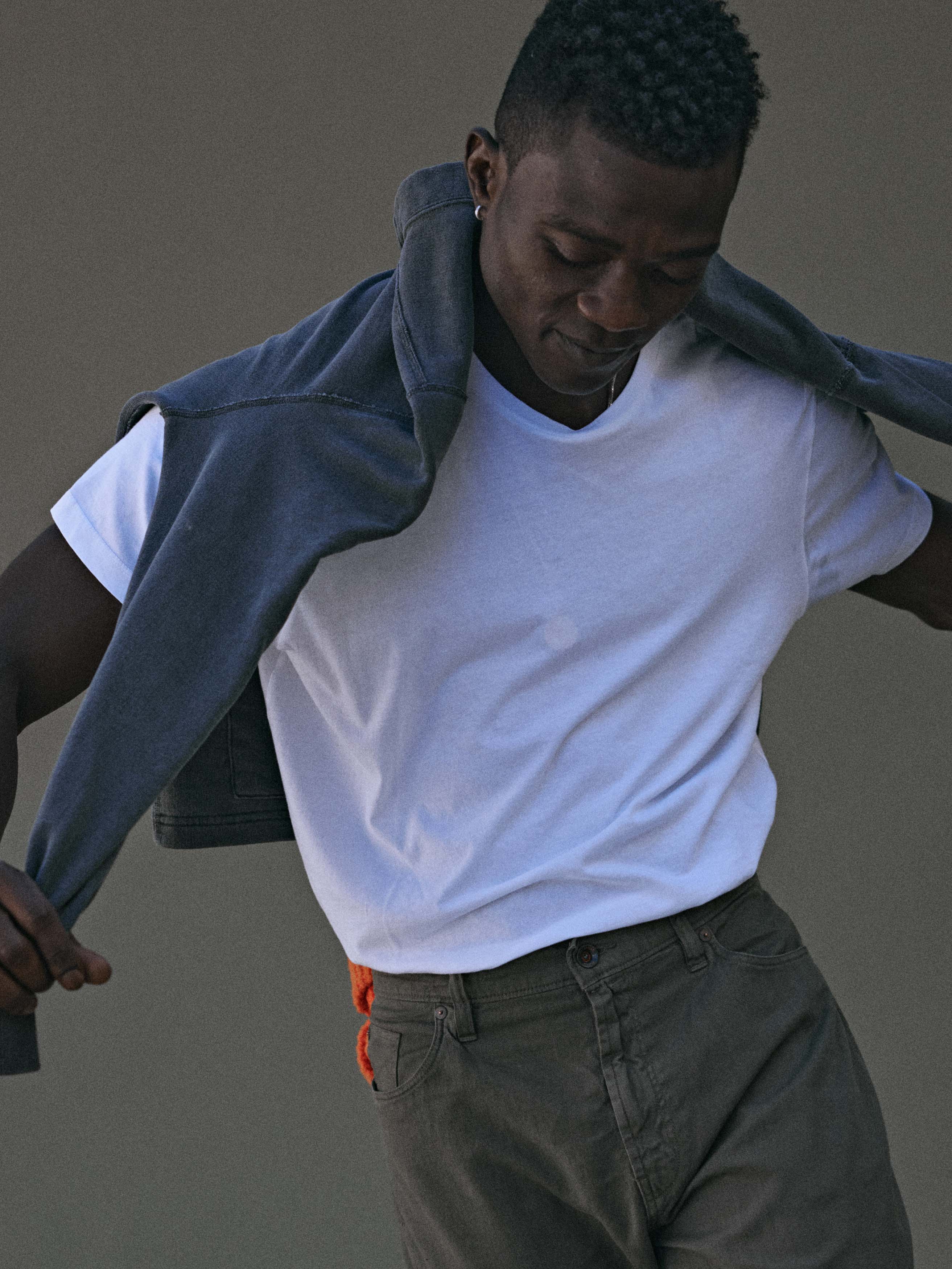 Portrait of a stylish dark-skinned adult male in a white t-shirt and olive-green pants with a grey sweater draped over his shoulders indoors.