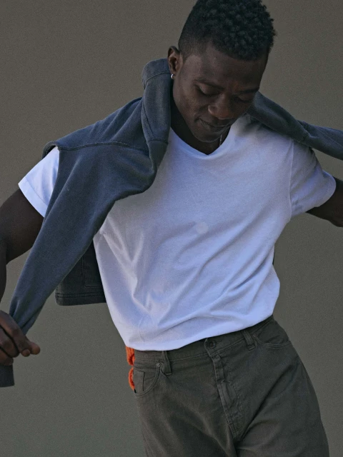 Portrait of a stylish dark-skinned adult male in a white t-shirt and olive-green pants with a grey sweater draped over his shoulders indoors.