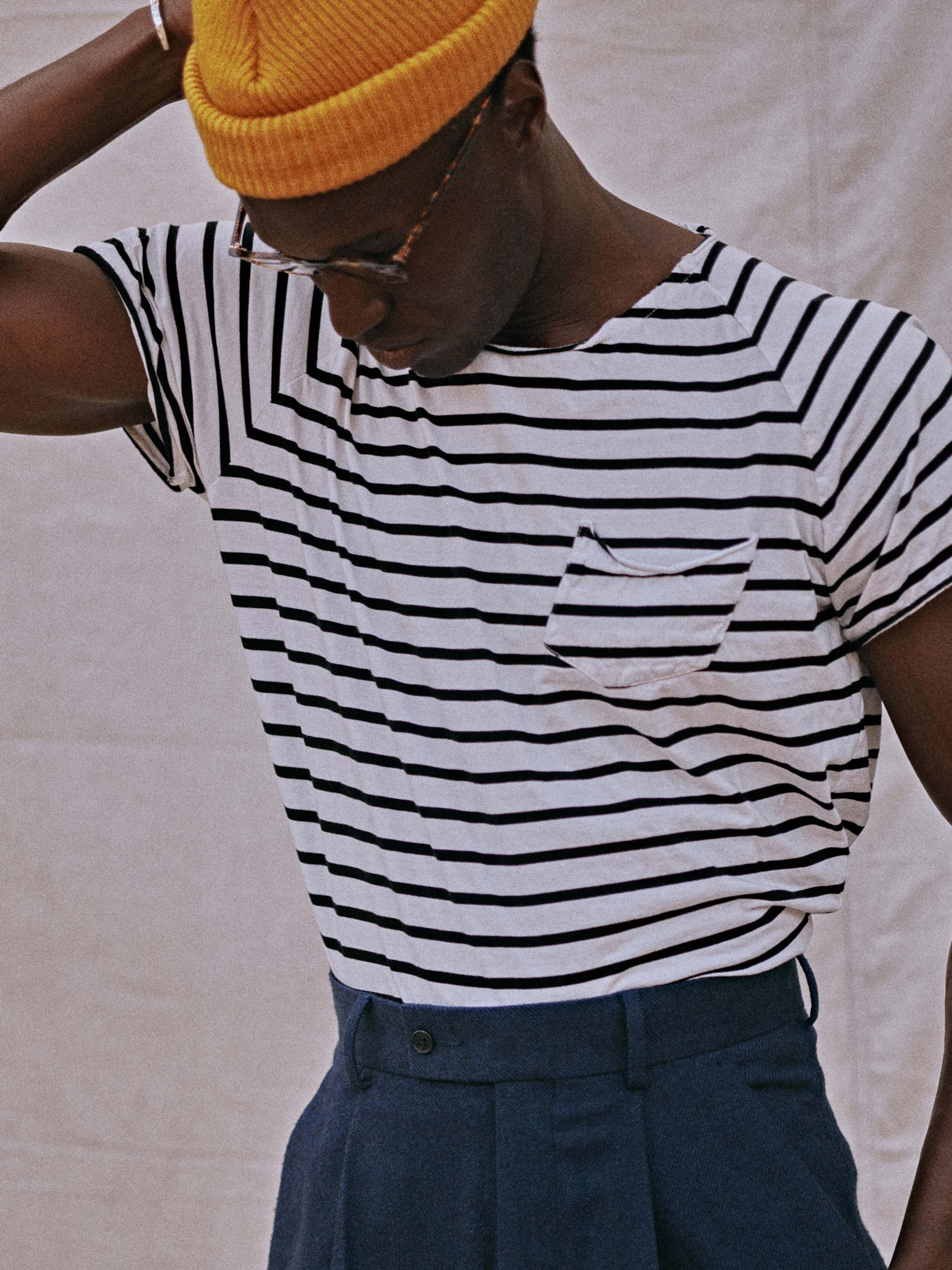 Young Black man poses in a striped shirt and blue pants, wearing a yellow beanie and glasses, against a neutral background.