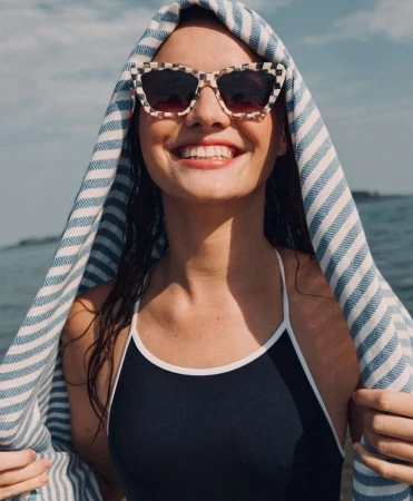 Smiling woman wearing checkered sunglasses with a blue and white striped towel draped over her head by the water
