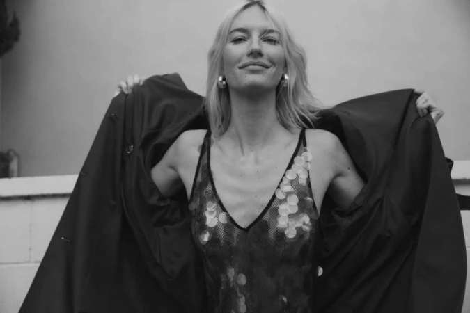 Black and white portrait of a smiling blonde woman wearing a sequined top and holding open a dark coat