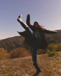 Woman in a long dark coat and boots kicking her leg up playfully in a grassy hillside landscape under a clear sky.