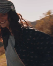 Smiling woman in a patterned cap and dark patterned jacket outdoors with motion blur captured by John Schell