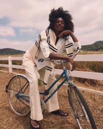 Black woman with dark curly hair wearing cream trousers and white shirt leaning on blue bicycle outdoors