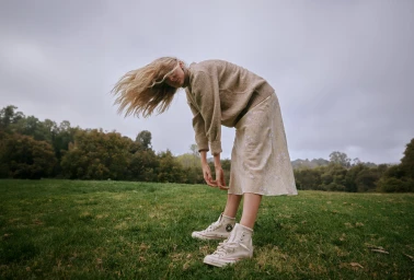 Blonde woman bending forward with hair whipping in the wind on a grassy field under an overcast sky.
