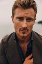 Close-up portrait of a man with blue eyes and stubble wearing a dark blazer over a blue button-down shirt outdoors.