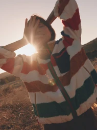 Model wearing a fuzzy multi-colored striped fleece jacket outdoors in a dry landscape during sunset.