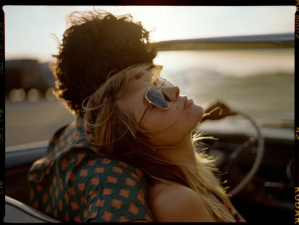 Close-up of a woman leaning on a man in a convertible car at sunset, wearing sunglasses and patterned shirt.