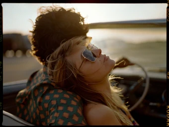 Close-up of a woman leaning on a man in a convertible car at sunset, wearing sunglasses and patterned shirt.