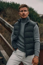 Man wearing a grey turtleneck sweater, dark puffer vest, and white jeans while posing near wooden steps outdoors.