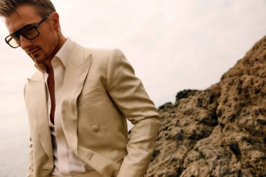 Man wearing a cream double-breasted suit and thick-rimmed glasses outdoors by rocky terrain, photographed by John Schell.