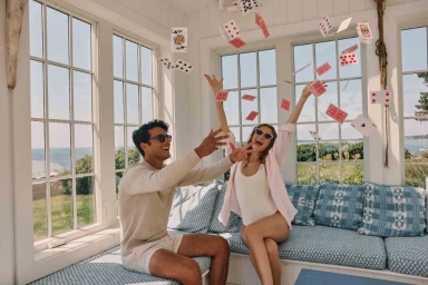 Man and woman throwing playing cards in the air inside a bright sunlit sunroom with large windows.