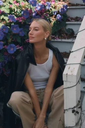 Blonde woman wearing white tank top and black leather jacket seated outdoors on outdoor stairs near purple flowers