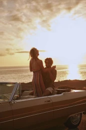 A smiling blonde woman stands in the back of a vintage convertible car next to a man at sunset by the ocean.