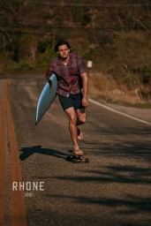 Man riding a skateboard on an asphalt road while carrying a surfboard under his arm.