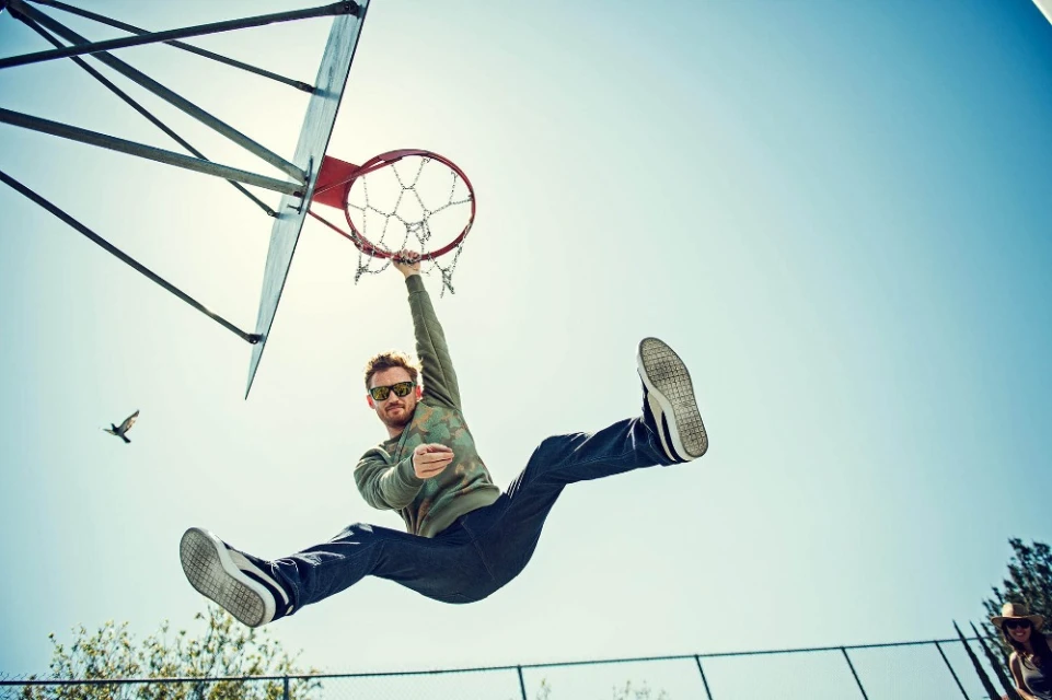 Adult male hangs from a basketball hoop in blue daylight sky while a bird flies by and a woman watches from a distance.