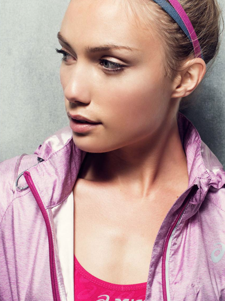 Close-up of a fair-skinned adult woman wearing a pink athletic jacket and headband, looking to the left against a grey backdrop.