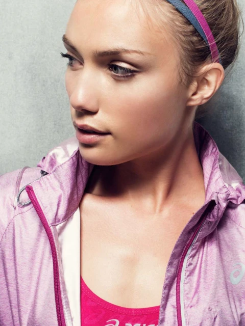 Close-up of a fair-skinned adult woman wearing a pink athletic jacket and headband, looking to the left against a grey backdrop.