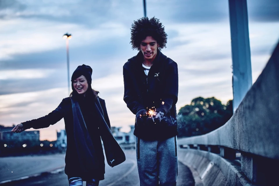Two people, an adult male and an adult female, are outside at dusk with sparklers, with a cityscape visible in the background.
