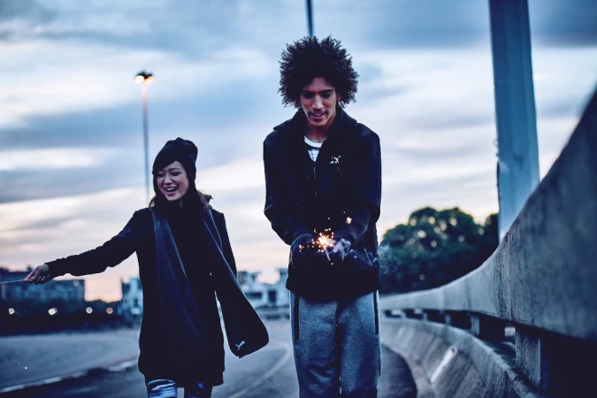 Two people, an adult male and an adult female, are outside at dusk with sparklers, with a cityscape visible in the background.