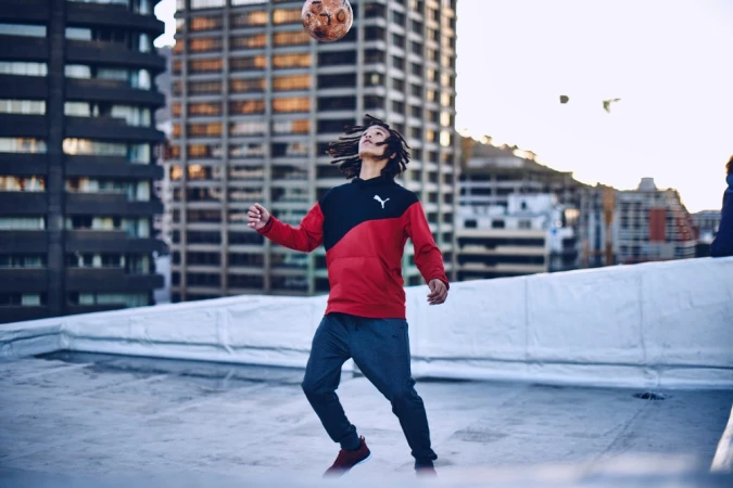 A man with dreadlocks juggles a soccer ball on a rooftop with a cityscape backdrop, wearing a Puma hoodie and sweatpants in an urban setting.