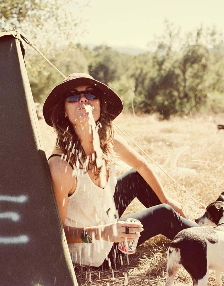 Woman outdoors in sunglasses spitting water, seated next to a tent, wearing a hat with a dog nearby on a sunny day.