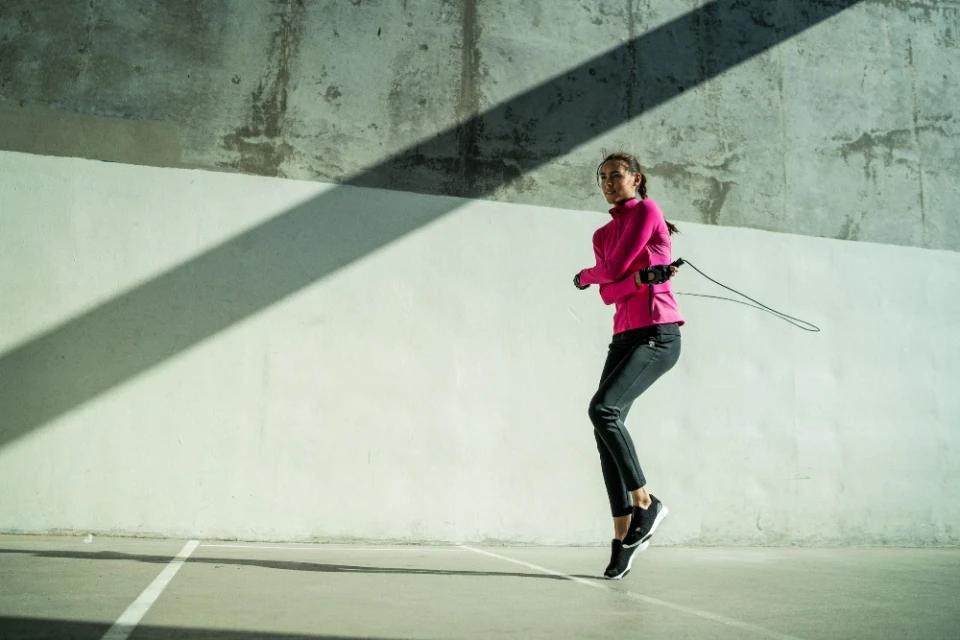Athletic woman in pink jacket jumps rope outdoors against a concrete wall with shadows. Workout, fitness, and active lifestyle themes.