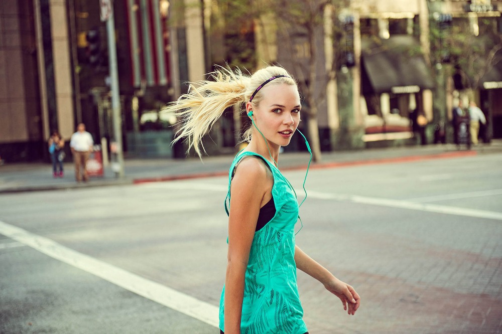 Blonde woman wearing a turquoise top walking down a city street with earbuds in