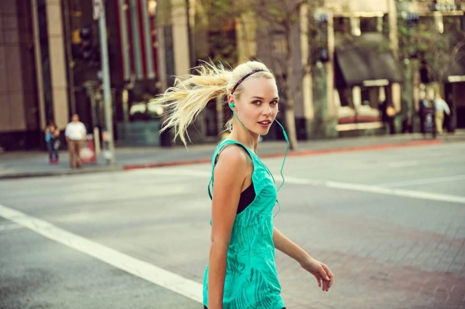 Blonde woman wearing a turquoise top walking down a city street with earbuds in