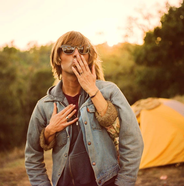 Outdoor portrait of a man blowing a kiss, wearing sunglasses and a denim jacket, with a woman's arm around him and a tent in the background.