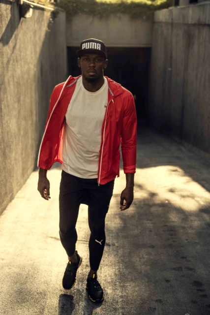 Athletic man in a red jacket and black pants walks towards the camera through an arched passage in the daytime. He is wearing a Puma hat.