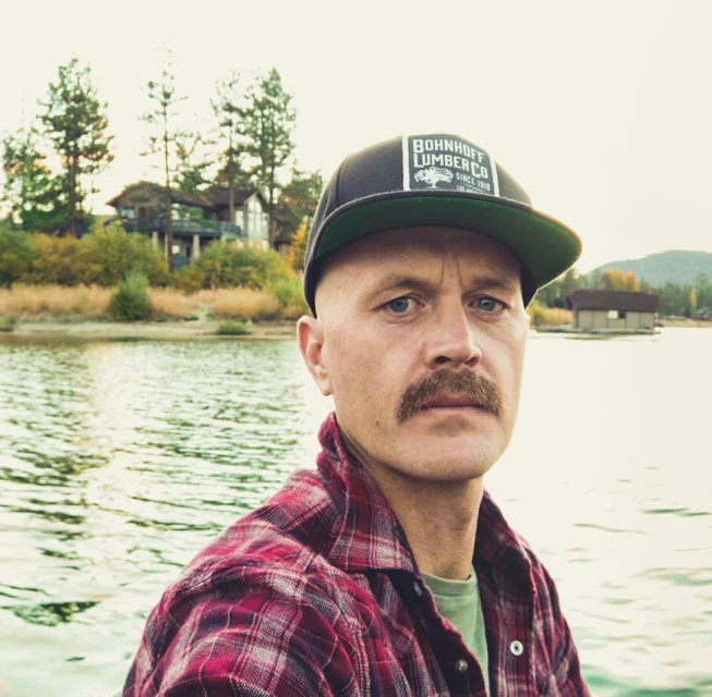 Outdoor portrait of a man with a mustache wearing a cap and flannel shirt, standing in front of a lake and trees in the background.