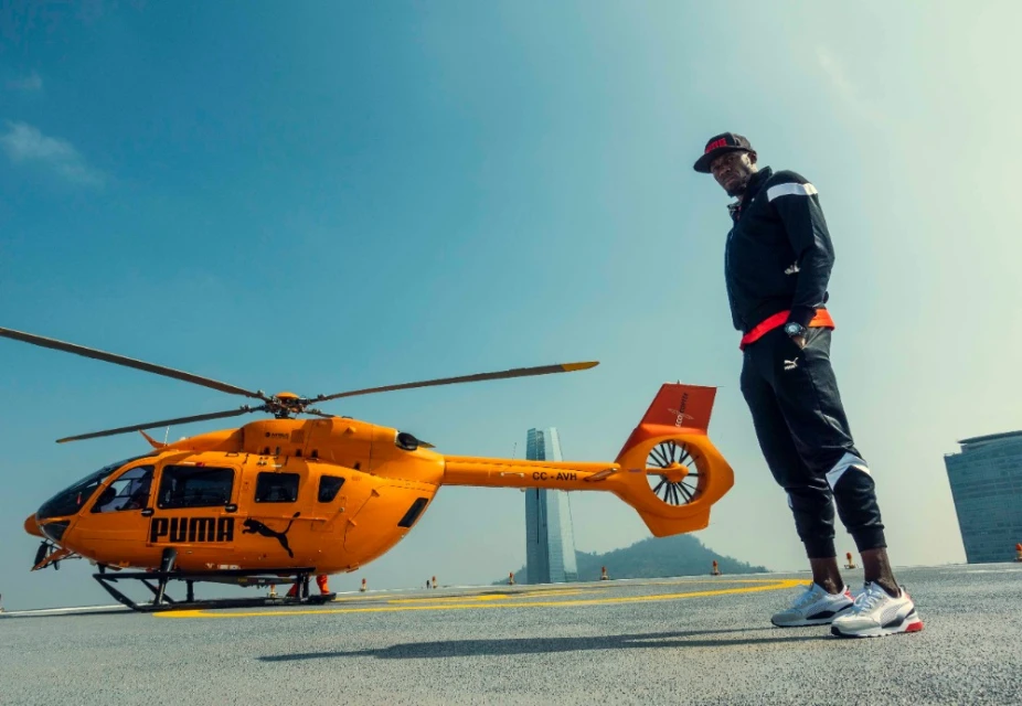 Athlete stands next to a yellow helicopter with Puma branding on a landing pad against a cityscape and bright blue sky.