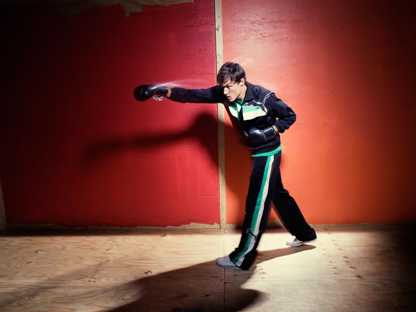 Young man in a tracksuit and boxing gloves throws a punch in a brightly lit, wood-floored room with orange and red walls.