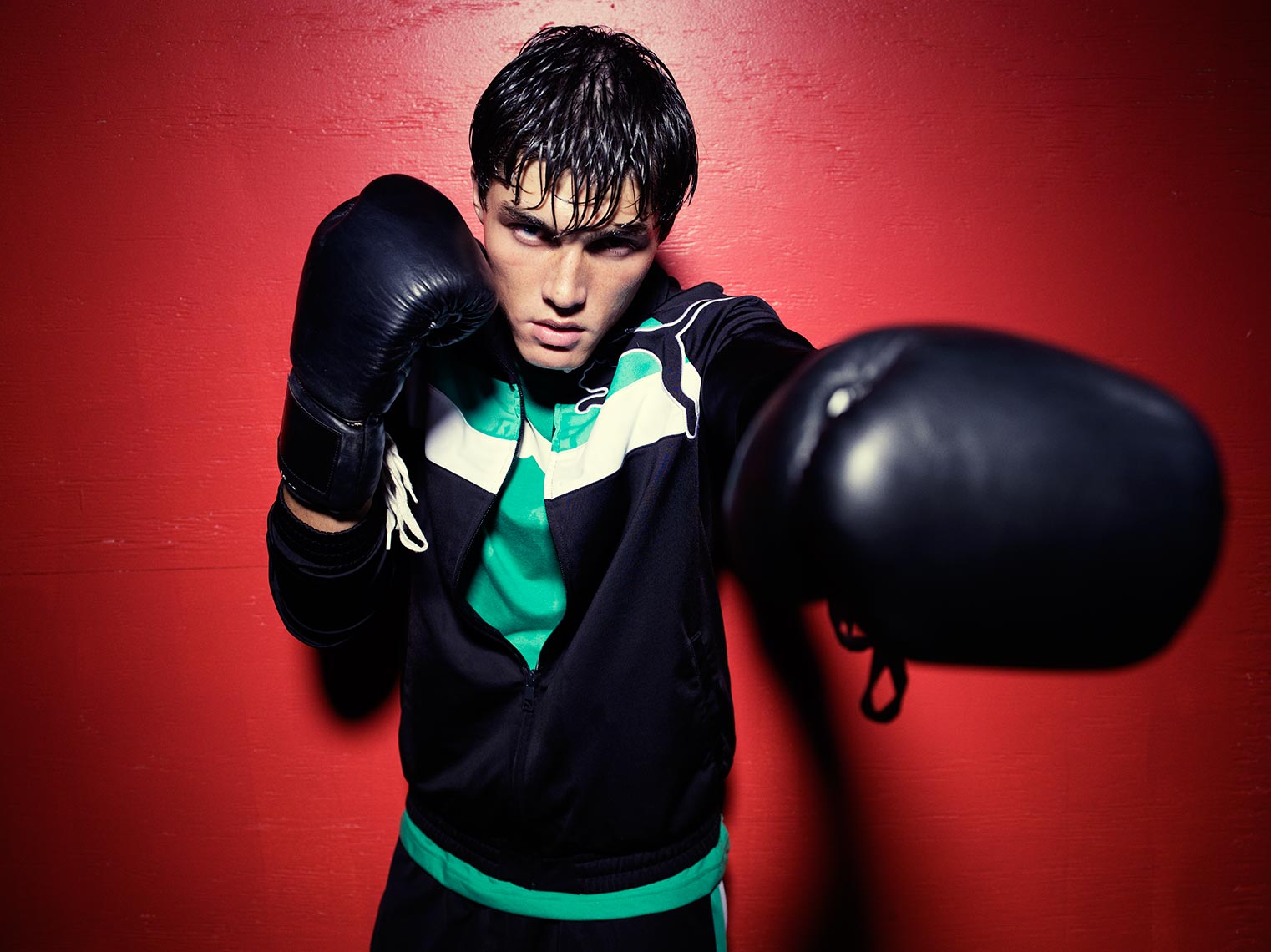 Young man in a black and green tracksuit and boxing gloves stands in front of a red wall, looking intensely at the camera.
