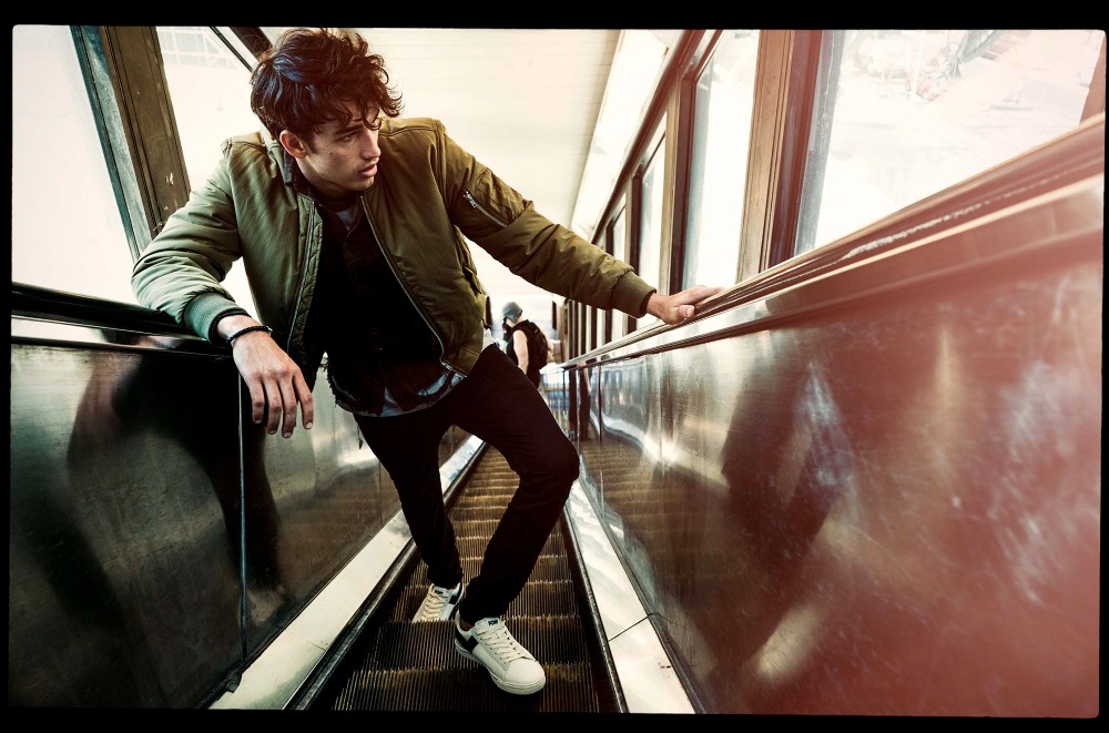 A young man on an escalator with a neutral expression, wearing a green jacket, black pants, and white sneakers, looking to the side.