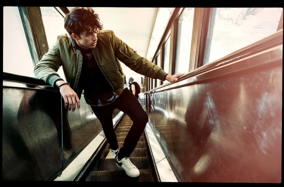 A young man on an escalator with a neutral expression, wearing a green jacket, black pants, and white sneakers, looking to the side.