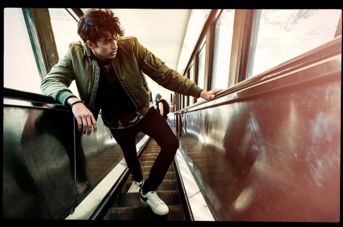 A young man on an escalator with a neutral expression, wearing a green jacket, black pants, and white sneakers, looking to the side.
