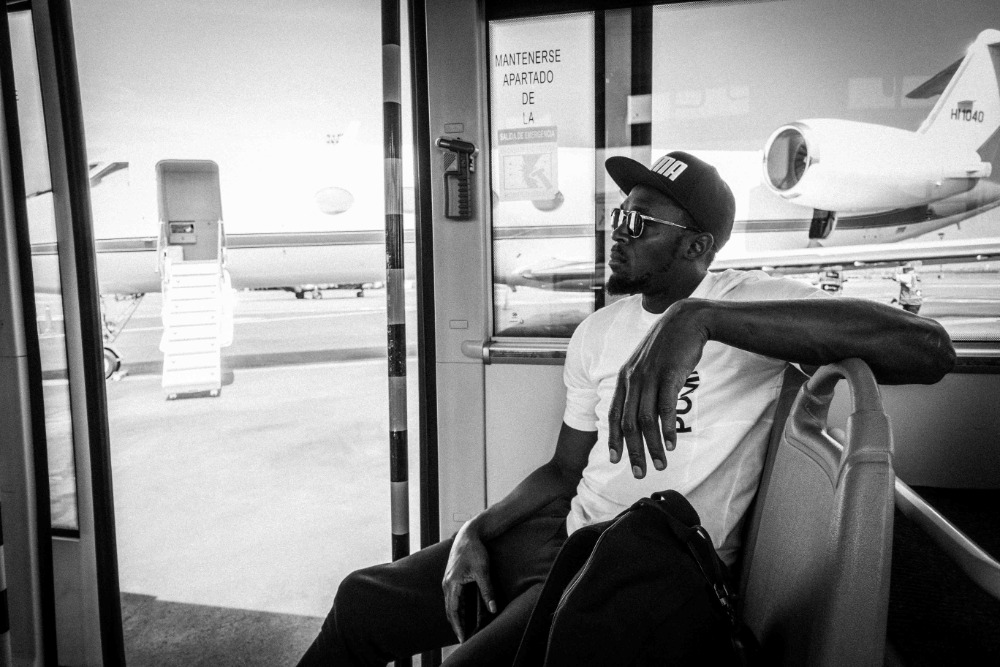 Monochrome image of an adult male in a cap and sunglasses sitting near an airplane, gazing out of a window on a moving bus at an airport.