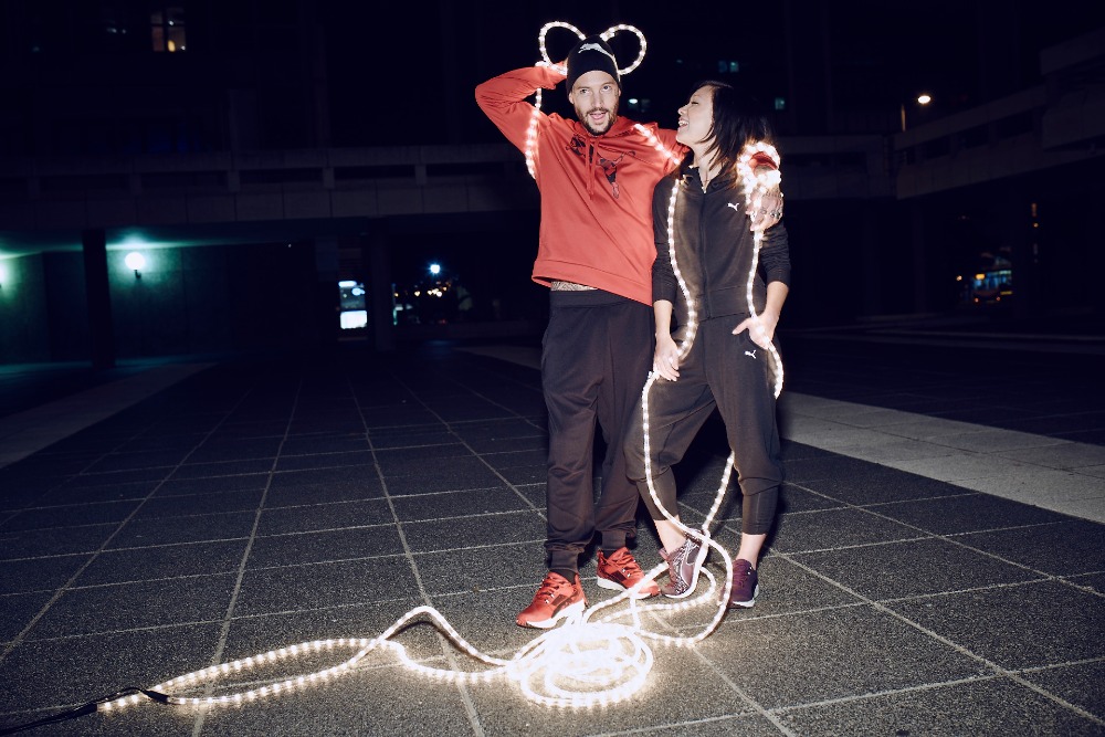 Two adults pose on a city plaza at night. Man in a red hoodie has light-up Mickey Mouse ears; woman in a black tracksuit with light trim.