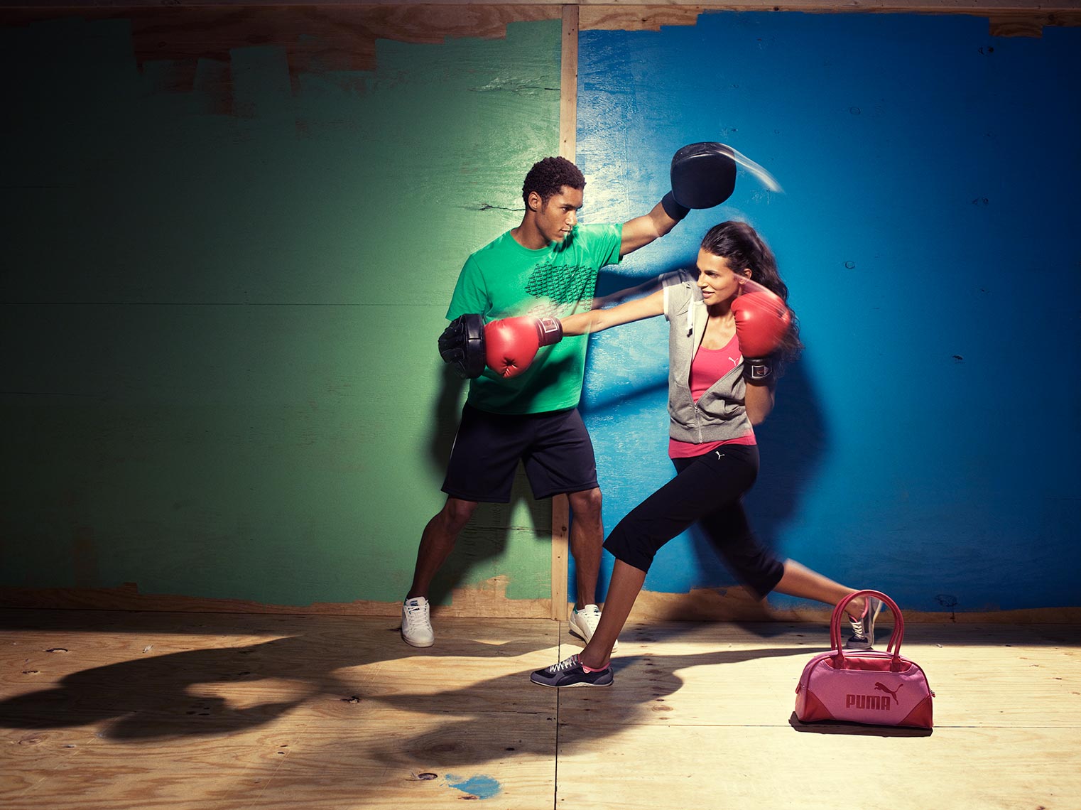 Man and woman boxing indoors. The male is wearing a green shirt and black shorts while the woman has on sportswear and boxing gloves.